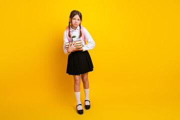 Young schoolgirl with a stack of book standing against vibrant yellow background, symbolizing...
