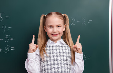 Smiling schoolgirl pointing up in classroom by the chalkboard with math equations in formal outfit