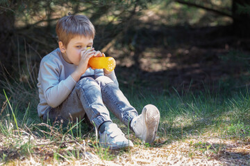 A little boy drinking juice on a hot summer day, in the forest, in the shade
