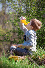 A little boy sits on the grass and greedily drinks orange juice