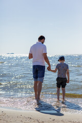 A father and son are wading in the water by the Baltic Sea, holding hands, and looking at the water.