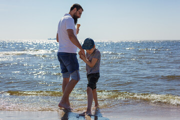 A bearded father and his little son are wading in the water and eating ice cream at the seaside