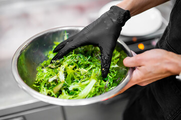 Chef wearing black gloves mixing fresh green salad in a stainless steel bowl, preparing healthy...
