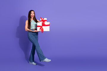 Young woman in casual attire smiling and holding gift boxes, standing against a vibrant purple...