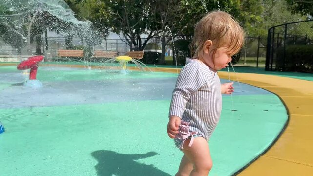 Cute Caucasian baby girl playing on a splash pad on a hot summer day. Baby playing with water outdoors. Water play kids