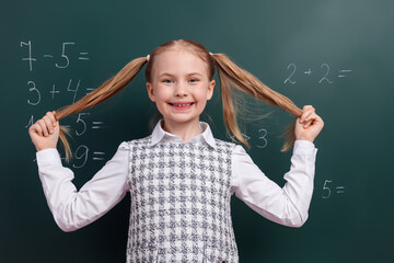 Cheerful young schoolgirl with ponytails posing in classroom against blackboard with math equations...