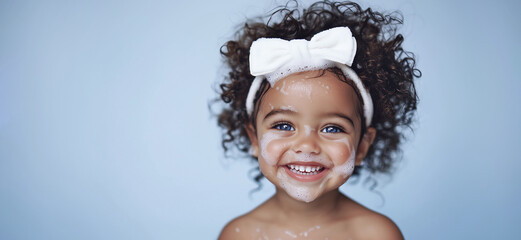 Portrait of cute smiling beautiful dark-skinned girl with blue eyes, in white headband on blue background with copy space. Happy child with wet hair, foam on head, hygiene and health