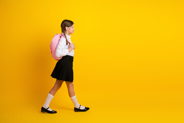 Cheerful schoolgirl walking with a pink backpack on a yellow background, expressing education and...