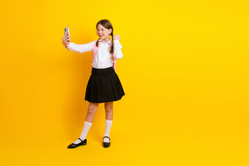 Happy schoolgirl in uniform taking a selfie with smartphone, standing on yellow background,...