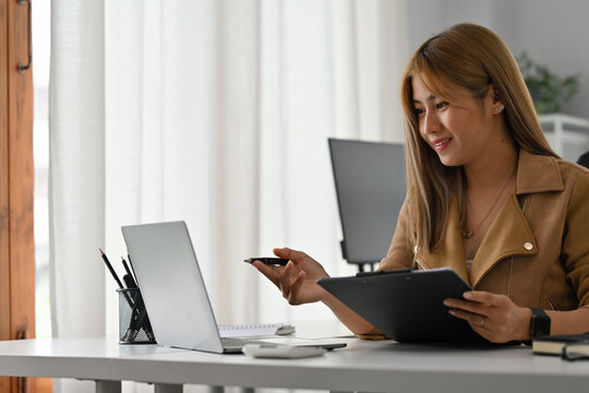 Smiling businesswoman holding a clipboard and pen during an online meeting via laptop in a modern office, Remote Business Meeting and Digital Collaboration