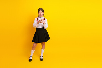 Smiling schoolgirl using a smartphone while standing against vibrant yellow background, capturing a...