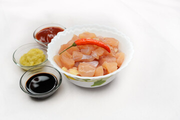 Raw chicken breast pieces in a white bowl with garlic and ginger paste, tomato ketchup, soya sauce, and other spices on a white background. Healthy food concept. Selective focus and copy space