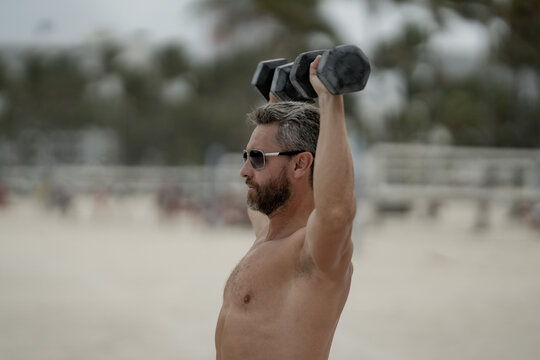 Sporty man lifting dumbbells outdoors. Sport man exercising with dumbbells in public outdoor gym. Fit guy working out in the park. Sportsman lifting dumbbells. 40s man training with dumbbells.