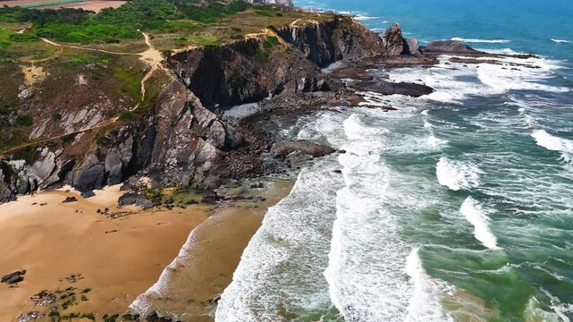 Wide aerial drone shot moving over large rolling waves and a rugged rocky beach with towering cliffs at Praia da Am&aacute;lia, Costa Vicentina, Portugal. Showcasing the powerful ocean meeting dramatic coast