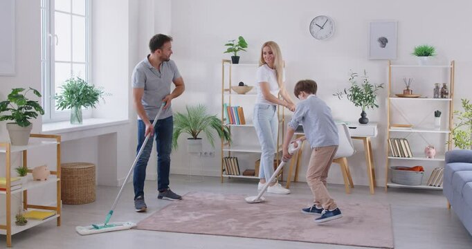 United happy family mother, father and son cleaning living room together with mop, vacuum cleaner and pp duster. Smiling young parents with little boy doing housework, housekeeping and household.