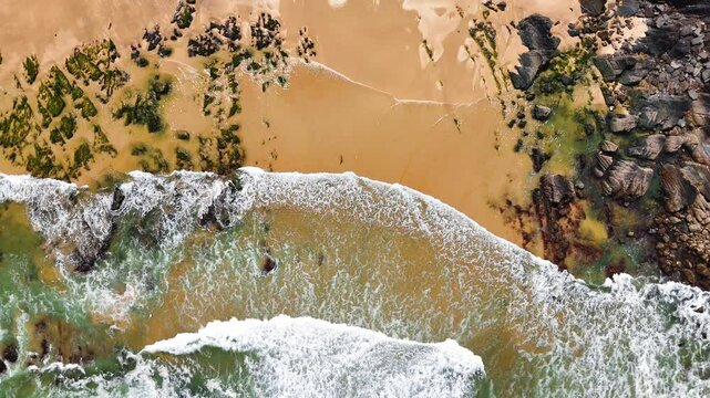 Top-down aerial view of ocean waves crashing onto the sand at Praia da Am&aacute;lia, Costa Vicentina, Portugal. Captured in slow motion, showing a dramatic coastal scene.