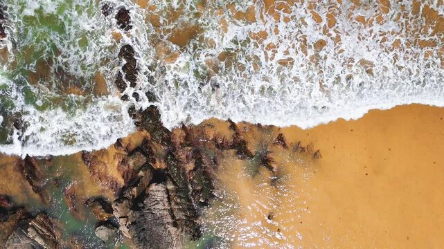 Top-down aerial view of ocean waves crashing onto the sand at Praia da Am&aacute;lia, Costa Vicentina, Portugal. Captured in slow motion, showing a dramatic coastal scene.
