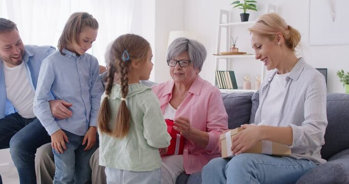 Two lovely children boy and girl congratulating their grandmother with birthday and giving her gift boxes. Happy big multi generation family sitting on sofa, celebrating special occasion together.