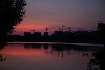 Construction cranes and urban buildings silhouette at sunset, reflected in lake with nature in foreground, evening skyline with industrial development. Contrast between nature and construction
