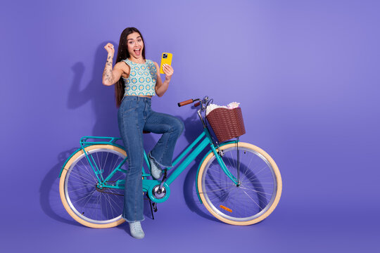 Happy young woman on a bicycle holding a smartphone against a vibrant purple background, celebrating with an excited hand gesture