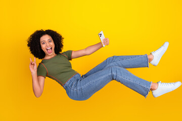Excited young woman posing against yellow background holding a phone, showing peace sign, and...
