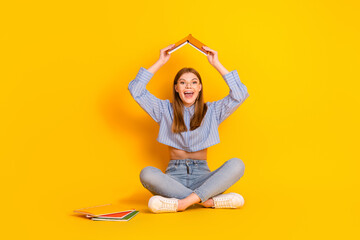 Cheerful young woman with a book on her head sitting on yellow background among scattered notebooks...