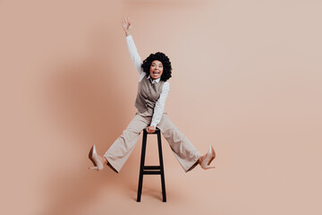 Elegant young businesswoman joyously posing in a stylish beige suit on a stool against a light...
