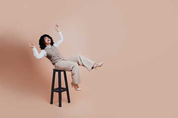 Gorgeous businesswoman in stylish attire gesturing enthusiastically on a high stool against a beige...