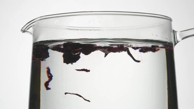 Hibiscus Falls Into the Kettle. Dried hibiscus petals slowly fall into a glass kettle with boiling water and stain the water pink