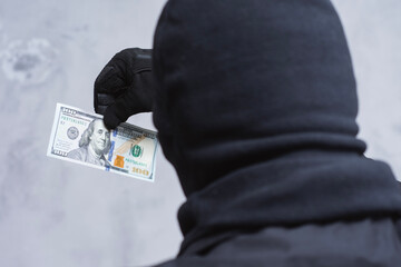 A mysterious figure dressed in a black mask and gloves holds a US hundred dollar bill. Concept...