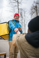 Delivery man wearing a thermal backpack, delivering a food order to a customer while using a mobile phone for contactless payment in an urban outdoor setting