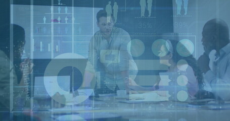 Man in light shirt leaning on table addressing team in office, with laptops and digital charts