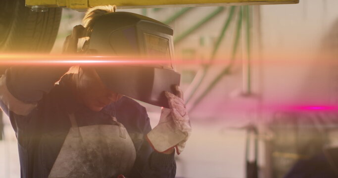Female technician lifting welding helmet from vehicle in repair workshop, with gloves and apron