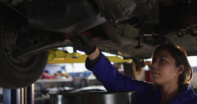 Female mechanic wearing blue coverall reaching under raised chassis in workshop, with oil drain pan