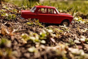 A bright red miniature toy car, a classic sedan model, sits on a patch of uneven, dry earth with...