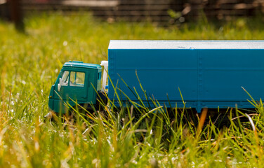 A miniature toy truck, green and blue, navigating through tall, sunlit green grass outdoors.