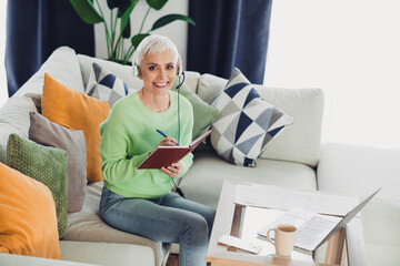 Obraz premium Confident woman with white hair enjoying leisure time at home, taking notes on a sofa with a laptop and a headset