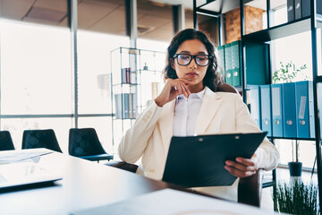 Professional businesswoman wearing formal suit reviewing documents in modern office workspace