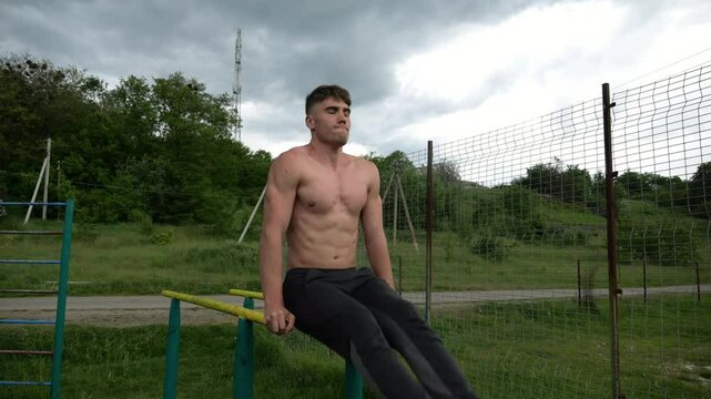 Strong man trains abdominal muscles raising legs on parallel bars at sports ground. Young shirtless sportsman exercising at nature. Sporty athlete working out at nature. Concept of sportive lifestyle