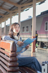 A young Asian woman stands on a public transport platform, holding a map, smiling slightly. She...