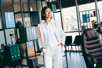 Confident businesswoman in white suit standing in modern corporate office with stylish interior and...