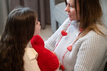 A mother and daughter share a tender moment, embracing with a red heart-shaped plush toy.