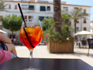 A refreshing Aperol Spritz cocktail garnished with orange slices sits on a café table in a sun-drenched Mediterranean plaza, perfect for evoking a summer holiday vibe.