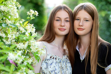 Two teenage sisters stand close together in a blooming garden, smiling softly. A touching portrait of sisterly love, trust, and emotional connection in nature