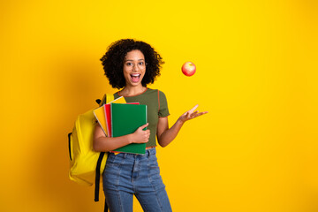 Cheerful student tossing apple with notebooks and backpack on vibrant yellow background, expressing casual school lifestyle