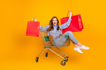 Cheerful young woman holding red shopping bags while sitting in a cart in a vivid yellow background