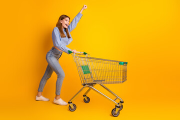 Excited young woman with shopping cart posing against yellow background highlighting casual style...