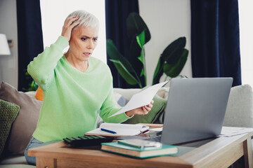 Concerned Senior Woman Reviewing Documents at Home with Laptop on Desk in Casual Living Room