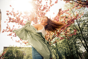 Young joyful woman enjoying a sunny day with cherry blossoms in an urban park setting, smiling with vibrant energy