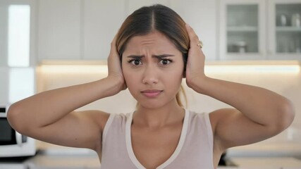 A young Asian woman with long brown hair covers her ears with her hands, showing a distressed expression in a modern kitchen setting.
